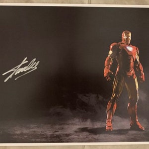 May include: A black and white poster featuring a red and gold Iron Man character standing in a smoky setting. The poster is signed "Stan Lee" in silver ink.