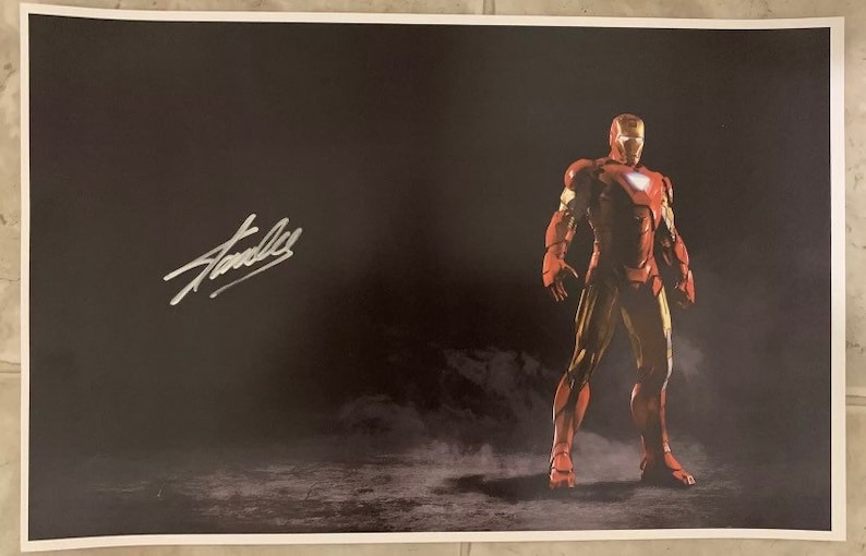 May include: A black and white poster featuring a red and gold Iron Man character standing in a smoky setting. The poster is signed "Stan Lee" in silver ink.