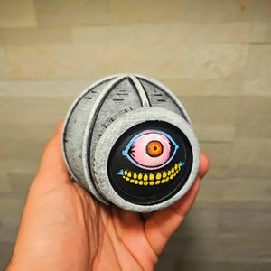 May include: A grey, spherical object featuring a cartoon eye design. The eye is pink with an orange iris, blue eyelids, and a yellow-toothed grin on a black background. The object has a textured, metallic appearance.