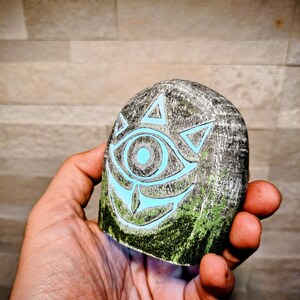 May include: A gray, hand-held stone with a carved design featuring a glowing blue eye and triangles. The bottom edge has green paint, and the stone is held against a textured background.