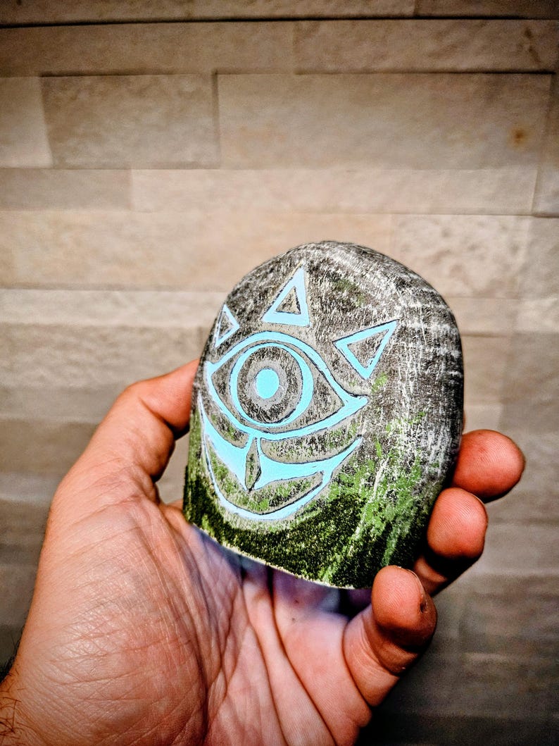 May include: A gray, hand-held stone with a carved design featuring a glowing blue eye and triangles. The bottom edge has green paint, and the stone is held against a textured background.
