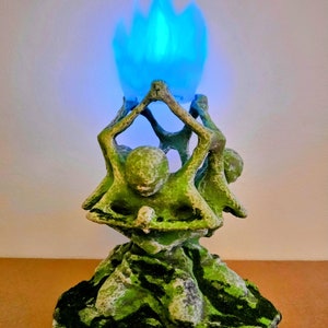 Puede incluir: Una escultura verde y musgosa de figuras sosteniendo una llama azul. Las figuras están en círculo, sosteniendo la llama. La base de la escultura tiene un aspecto texturizado y musgoso. La escultura tiene el texto "トーティフォス KNIGHTS" en la base.