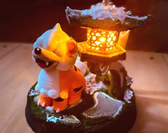 May include: A 3D-printed, illuminated sculpture featuring an orange and white creature next to a Japanese-style lantern. The lantern has a warm, glowing light. The base is dark with green moss and a stone.