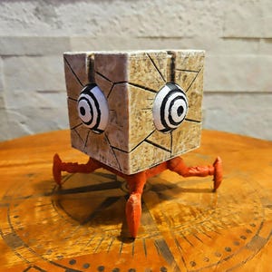 Może przedstawiać: Kapryśna rzeźba w kształcie kostki z teksturowanym, jasnobrązowym zewnętrzem. Posiada dwa duże, stylizowane oczy z czarno-białymi okręgami koncentrycznymi. Rzeźba wsparta jest na czterech pomarańczowych nogach.