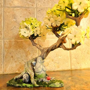 May include: A decorative bonsai tree sculpture featuring a seated Buddha figure. The tree has a textured brown trunk and branches with yellow and white flowers. A small red apple sits near the Buddha. The base is green and mossy.