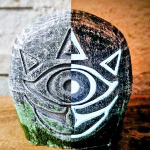 May include: A stone sculpture split vertically, with the left side in shades of gray and the right in brown. The sculpture features a stylized eye design with white accents and triangular shapes, and a base of green moss.