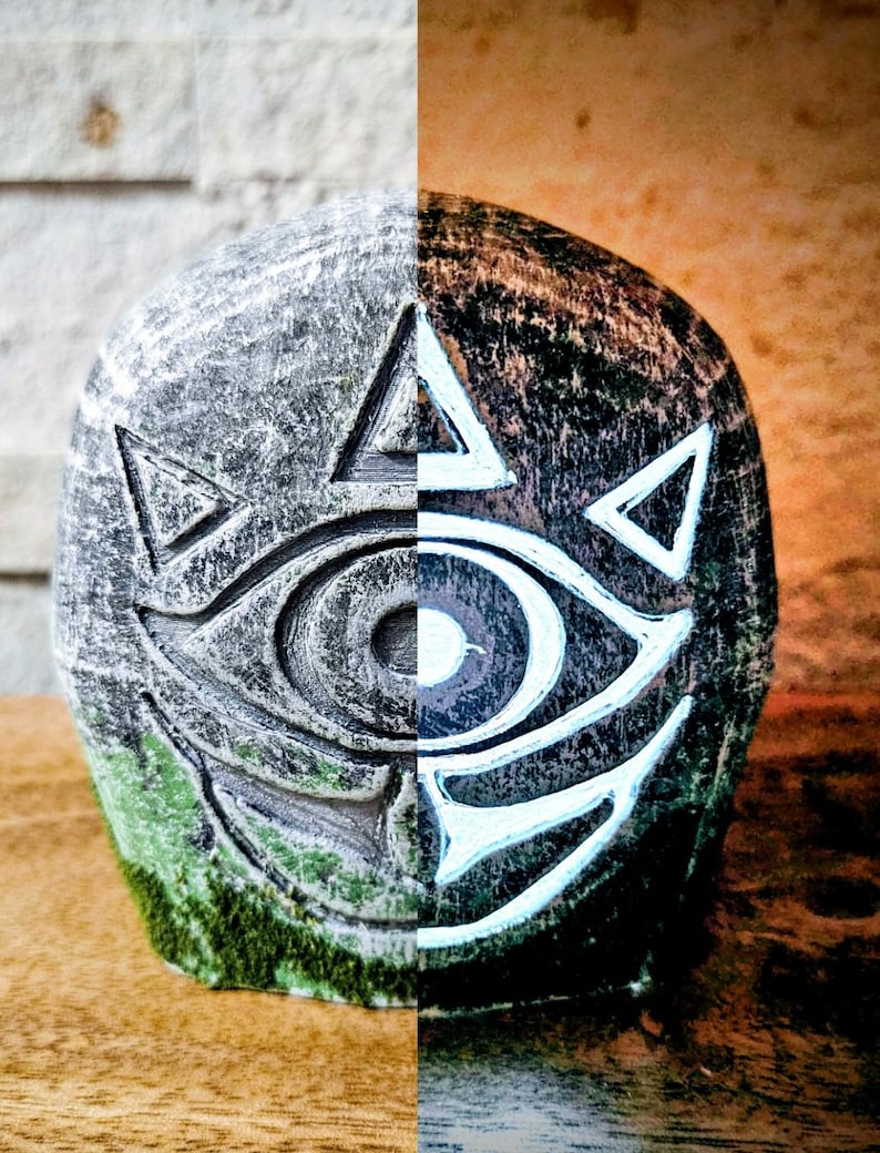 May include: A stone sculpture split vertically, with the left side in shades of gray and the right in brown. The sculpture features a stylized eye design with white accents and triangular shapes, and a base of green moss.