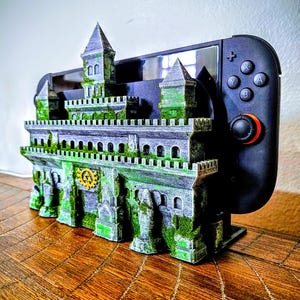 May include: A 3D-printed castle-shaped gaming console stand in gray, green, and black. The stand features a detailed castle design with turrets, walls, and a gold emblem. A black gaming controller is inserted into the stand.