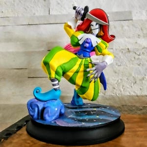 May include: A vibrant figurine of a jester, featuring a red hat, a yellow and green striped costume, and blue footwear. The jester stands on a starry base with a black edge. The figure has a white mask and holds a small item.