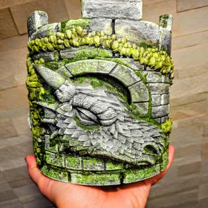May include: A cylindrical, gray and green dragon-themed sculpture. The sculpture features a dragon's head with detailed scales and a horn, integrated into a castle tower design. Green moss-like details add texture.