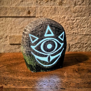 May include: A dark gray, rounded stone sculpture featuring a glowing blue eye design. The eye is surrounded by triangles and a curved line, with a detailed iris. The sculpture sits on a wooden surface.