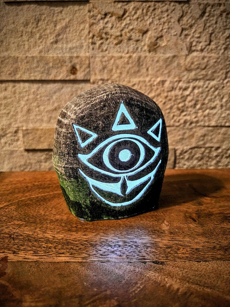 May include: A dark gray, rounded stone sculpture featuring a glowing blue eye design. The eye is surrounded by triangles and a curved line, with a detailed iris. The sculpture sits on a wooden surface.