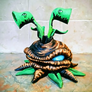 May include: A quirky, hand-painted sculpture of a plant-like creature. The sculpture has a brown and black textured body with blue eyes and green, leaf-like appendages. Two green stalks with eye-like details extend upwards.