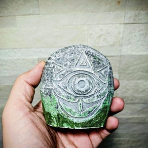 May include: A gray stone carving of an eye with a triangle above it and a smiling mouth below. The bottom of the stone has green moss. The carving is held in a hand.