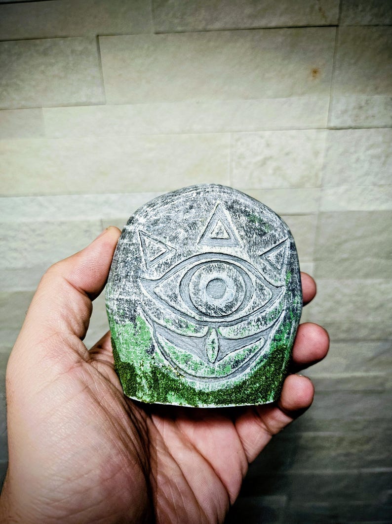 May include: A gray stone carving of an eye with a triangle above it and a smiling mouth below. The bottom of the stone has green moss. The carving is held in a hand.