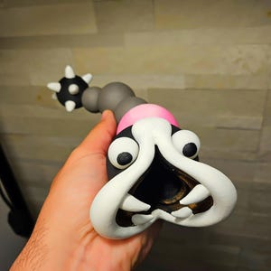 May include: A whimsical, handmade sculpture of a cartoonish creature. The sculpture features a black head with large white teeth and eyes, a pink collar, and a gray body. The tail end has a black ball with white spikes.