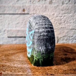 May include: A hand-painted, gray stone sculpture with a teal geometric design on the side. The bottom of the stone has a green moss-like texture. The sculpture is displayed on a wooden surface with a textured brick background.