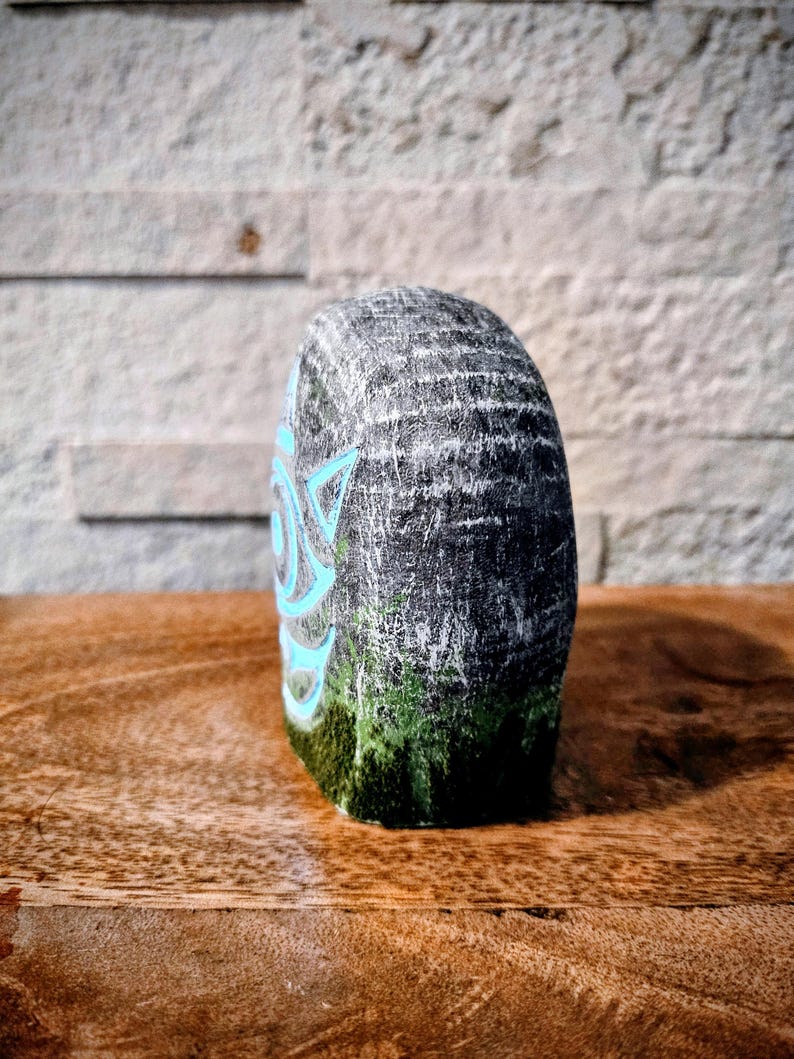 May include: A hand-painted, gray stone sculpture with a teal geometric design on the side. The bottom of the stone has a green moss-like texture. The sculpture is displayed on a wooden surface with a textured brick background.