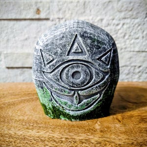 May include: A gray stone sculpture with an eye and triangle design. The sculpture has a weathered appearance with green moss at the base. The object is set on a wooden surface.