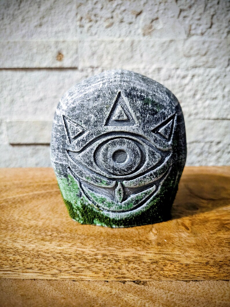 May include: A gray stone sculpture with an eye and triangle design. The sculpture has a weathered appearance with green moss at the base. The object is set on a wooden surface.