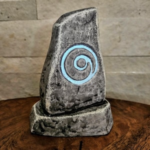 May include: A gray stone sculpture with a blue spiral design. The sculpture has a rough, textured surface and sits on a matching base. The background is a light-colored, textured wall.