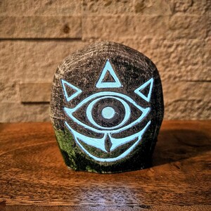May include: A dark, textured stone sculpture featuring a glowing teal eye design with triangular accents. The eye is the focal point, set against a dark, mottled background. The sculpture sits on a wooden surface.