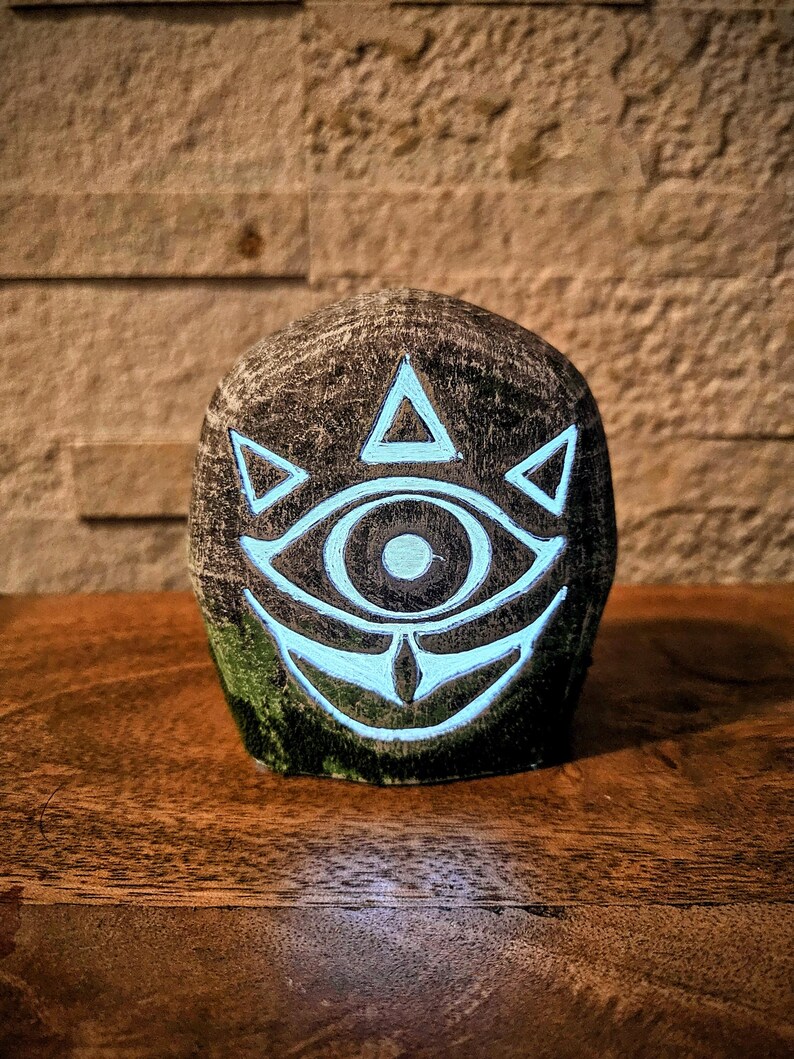 May include: A dark, textured stone sculpture featuring a glowing teal eye design with triangular accents. The eye is the focal point, set against a dark, mottled background. The sculpture sits on a wooden surface.