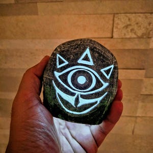 May include: A hand-painted, dark gray stone featuring a glowing white eye and triangle design. The stone has a rough texture and is held against a warm, neutral background. The design is reminiscent of a video game.