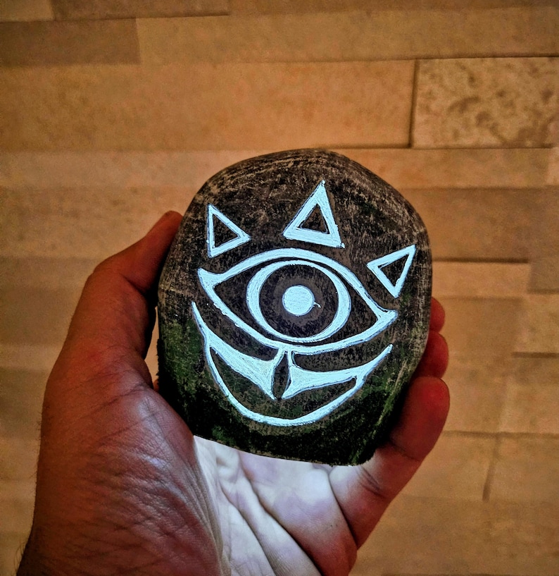 May include: A hand-painted, dark gray stone featuring a glowing white eye and triangle design. The stone has a rough texture and is held against a warm, neutral background. The design is reminiscent of a video game.