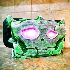 May include: A green and gray decorative case with a skull design, featuring two large pink eye sockets. The case is designed to fit a black handheld gaming console, with the console's buttons visible on the left side. The case has a textured, weathered appearance.