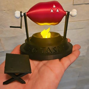 May include: A 3D printed model of a rotisserie chicken with a red chicken leg and a black base with the words "SO TASTY" printed on it. The chicken is rotating on a spit over a small fire.