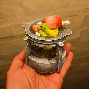 Könnte beinhalten: Ein Miniatur-Metallherd mit einem roten und grünen Lebensmittel darauf. Der Herd hat eine graue und weiße Lackierung.