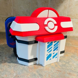 Puede incluir: Una funda multicolor impresa en 3D diseñada para parecerse a un edificio. La funda presenta un techo rojo con detalles blancos, una fachada blanca y detalles de ventanas azules y blancas. Un mando de juego azul y negro está adjunto al lateral.