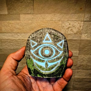May include: A hand holding a stone carved with a glowing blue eye design. The stone has a textured, gray surface with green accents. The eye symbol is surrounded by triangles and a curved line.