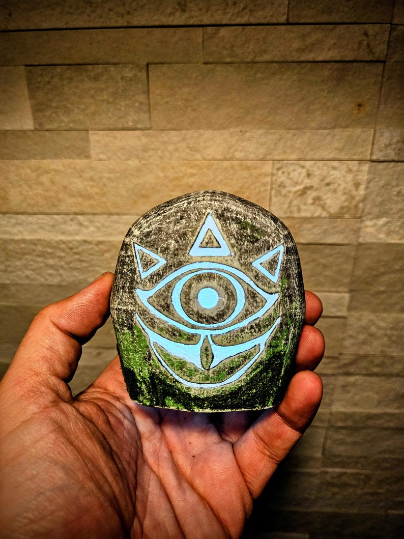 May include: A hand holding a stone carved with a glowing blue eye design. The stone has a textured, gray surface with green accents. The eye symbol is surrounded by triangles and a curved line.