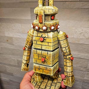 Peut inclure: Figurine de robot jaune doré avec une texture en forme de briques. Le robot a des yeux rouges, des accents circulaires rouges et des détails blancs en forme d'étoile. Un objet décoratif unique et artisanal.