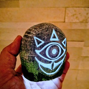 May include: A hand-carved, rounded stone featuring a glowing white eye design with triangular accents. The stone has a textured, dark gray surface with patches of green moss. The design is reminiscent of a video game.