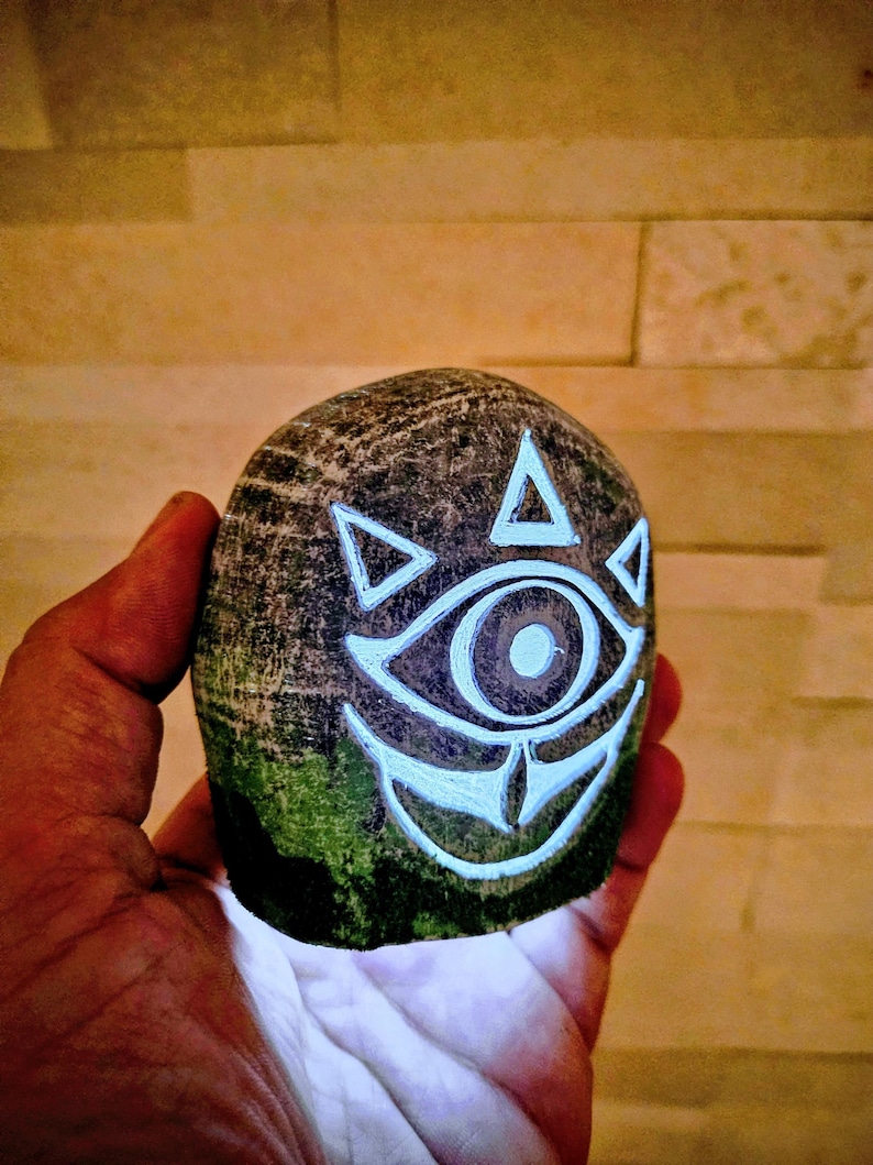 May include: A hand-carved, rounded stone featuring a glowing white eye design with triangular accents. The stone has a textured, dark gray surface with patches of green moss. The design is reminiscent of a video game.