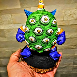 May include: A green, multi-eyed creature figurine with blue accents and a striped horn. The figurine is mounted on a black base with a wooden edge. The creature's eyes are yellow and white.