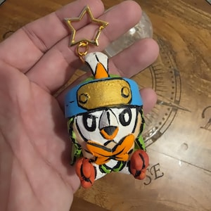 May include: A handcrafted keyring featuring a small, sculpted character with a white face, orange beak, and orange feet. The figure wears a blue headband with a gold-coloured plate and a bright orange bow tie, attached to a gold-coloured star-shaped clasp.
