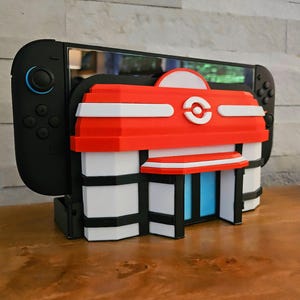 Peut inclure: Un étui décoratif rouge, blanc et noir pour une Nintendo Switch. L'étui est conçu pour ressembler à un bâtiment avec un emblème circulaire. La Switch est noire avec des boutons bleus et gris. L'étui est sur une surface en bois.