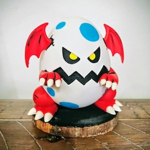 May include: A white egg-shaped figurine with blue spots, red wings, and red feet with white claws. The egg has a jagged black mouth and yellow eyes. It is mounted on a black base atop a wooden slice.