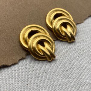 May include: A pair of gold-toned earrings with a knotted design. The earrings are made of a smooth, shiny metal.