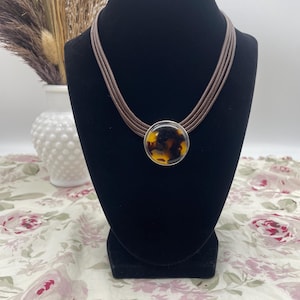 May include: A multi-strand brown cord necklace featuring a circular pendant with a polished silver-tone rim and a mottled amber and black tortoiseshell-patterned center. The necklace is displayed on a black velvet-covered neck form.