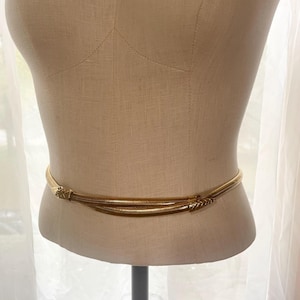 May include: A gold-toned metal snake belt featuring a flexible, coiled body design. The belt ends in detailed, textured snake head and tail motifs that meet at the front, creating a continuous, wrap-around appearance with a polished finish.