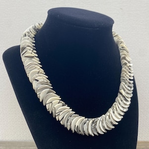 Op de afbeelding: Een opvallende ketting bestaande uit talrijke waaiervormige, crèmekleurige en grijze elementen. De ketting wordt gepresenteerd op een zwarte mannequin, die de gebogen vorm en het gedetailleerde ontwerp benadrukt. Een uniek accessoire.