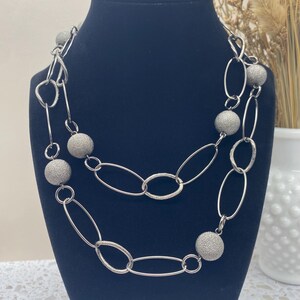 以下が含まれることがあります： テクスチャ加工されたシルバーボールが付いたシルバーチェーンネックレス。ネックレスは重ねてあり、楕円形のリンクがあります。