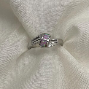 May include: A silver ring with two rectangular, iridescent opal stones. The ring band has a split design with a subtle, geometric pattern. The opals display a mix of pink, green, and purple hues, set against a white background.