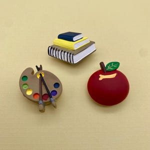 Pode incluir: Três puxadores de gaveta de cerâmica coloridos, um com a forma de uma pilha de livros, outro com a forma de uma paleta de pintura com pincéis e outro com a forma de uma maçã vermelha.