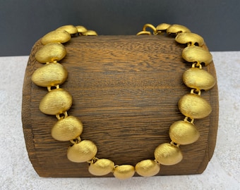 May include: A gold necklace made of round, brushed metal links. The necklace is displayed on a wooden stand.
